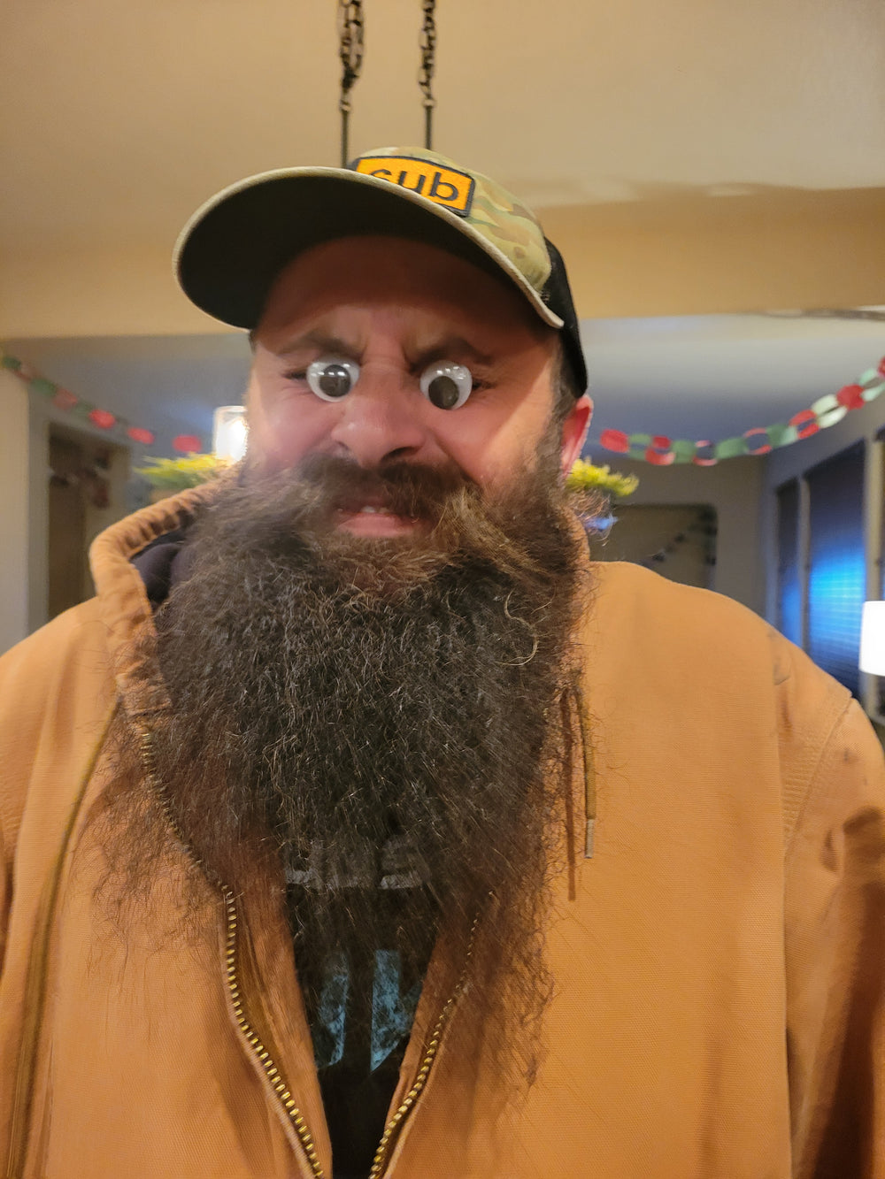A close-up portrait of a man with a long gray beard wearing a tan zip-up jacket and a camouflage baseball cap with yellow lettering, photographed indoors with a tilted camera angle.
