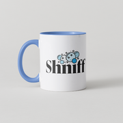 A white ceramic coffee mug with a blue handle and blue interior, featuring the word 'Shniff' in black stylized text with a blue cartoon sniffer graphic above it, on a light gray background.