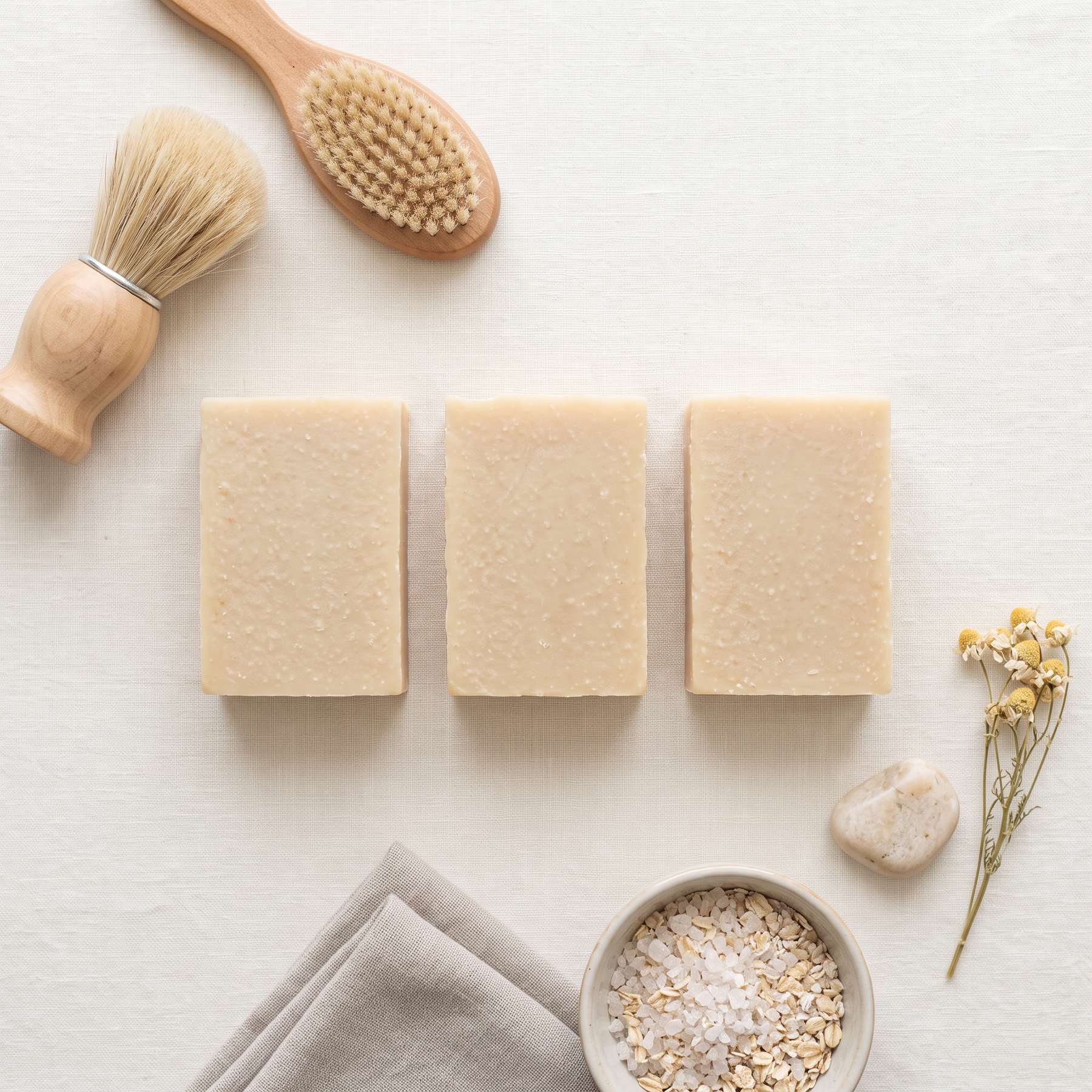 Three rectangular bars of pale beige soap arranged on a light neutral surface, styled with a wooden brush, wooden spoon, folded cloth, oatmeal bowl, and small dried flowers.